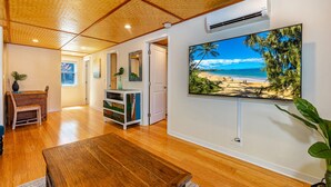 A spacious living room with hardwood flooring, a wooden ceiling, a large flat-screen TV mounted on the wall, and a view of a beach through the window. - Two homes w/ pool and hot tub near 3 beaches, Kihei (Kihei)