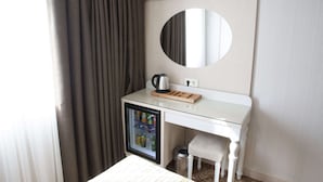 Habitación de hotel compacta con un escritorio pequeño, un refrigerador, un espejo y una ventana con cortinas.