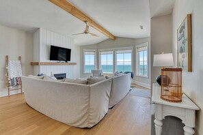 Uma sala de estar com um amplo sofá em formato de L, uma TV de tela plana, uma lareira e vista para o oceano.