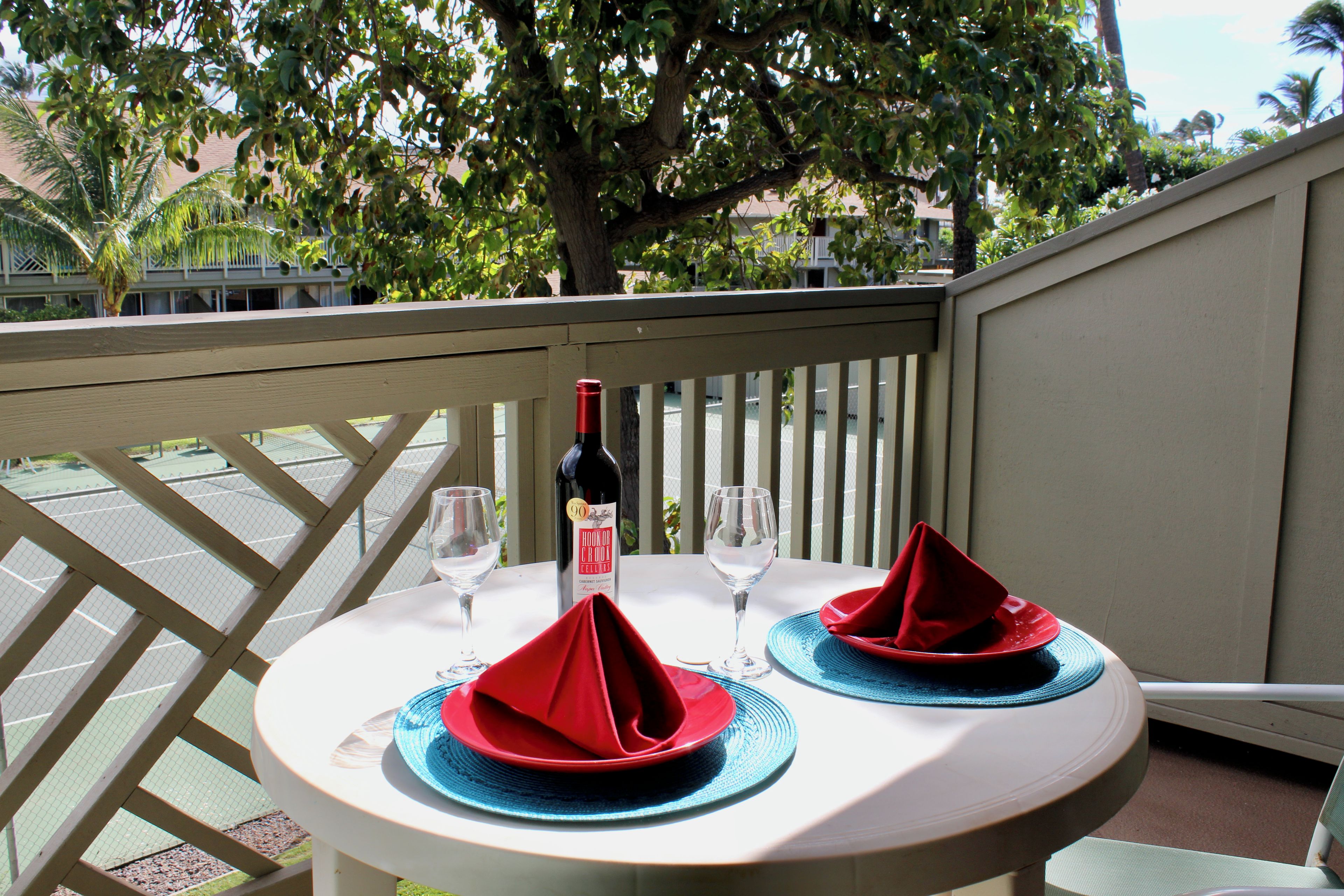 Een tafel voor twee, gedekt met rode servetten en blauwe placemats, een fles wijn en twee wijnglazen op een balkon met uitzicht op bomen en gebouwen.