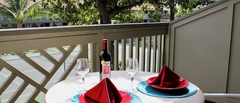 A table set for two with red napkins and blue placemats, a bottle of wine, and two wine glasses on a balcony with a view of trees and buildings.