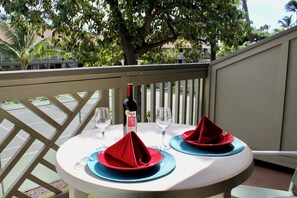 A table set for two with red napkins and blue placemats, a bottle of wine, and two wine glasses on a balcony with a view of trees and buildings. - Kihei Bay Surf #218 Renovated, Near Beach, Shared Hot Tub, Inner Courtyard (Kihei)