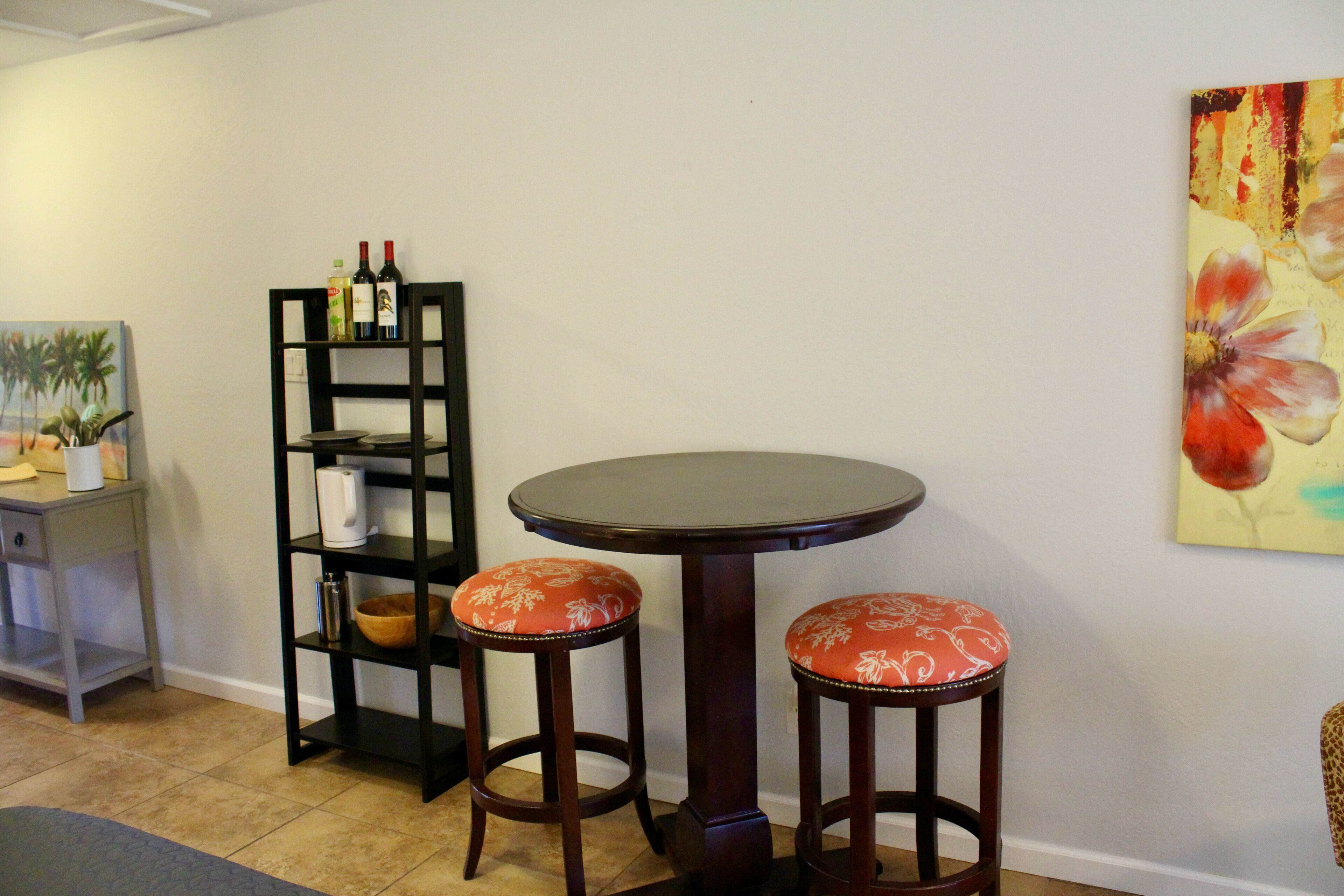 A room with a round table and two stools with floral upholstery, a tall black bookshelf with wine bottles, and a painting on the wall.