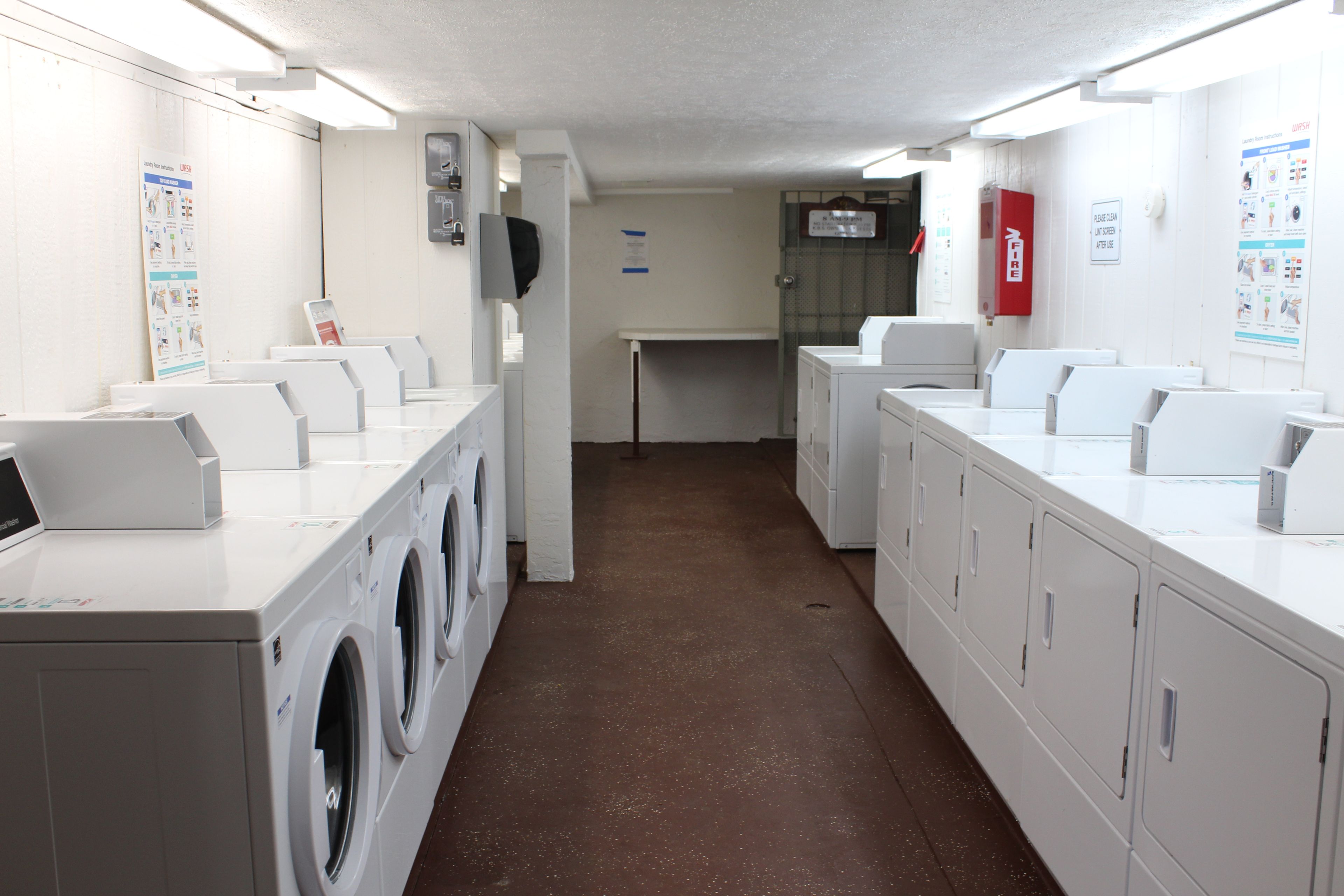 Une buanderie équipée de plusieurs machines à laver et de plusieurs sèche-linge, d’un extincteur et d’affiches informatives accrochées aux murs.