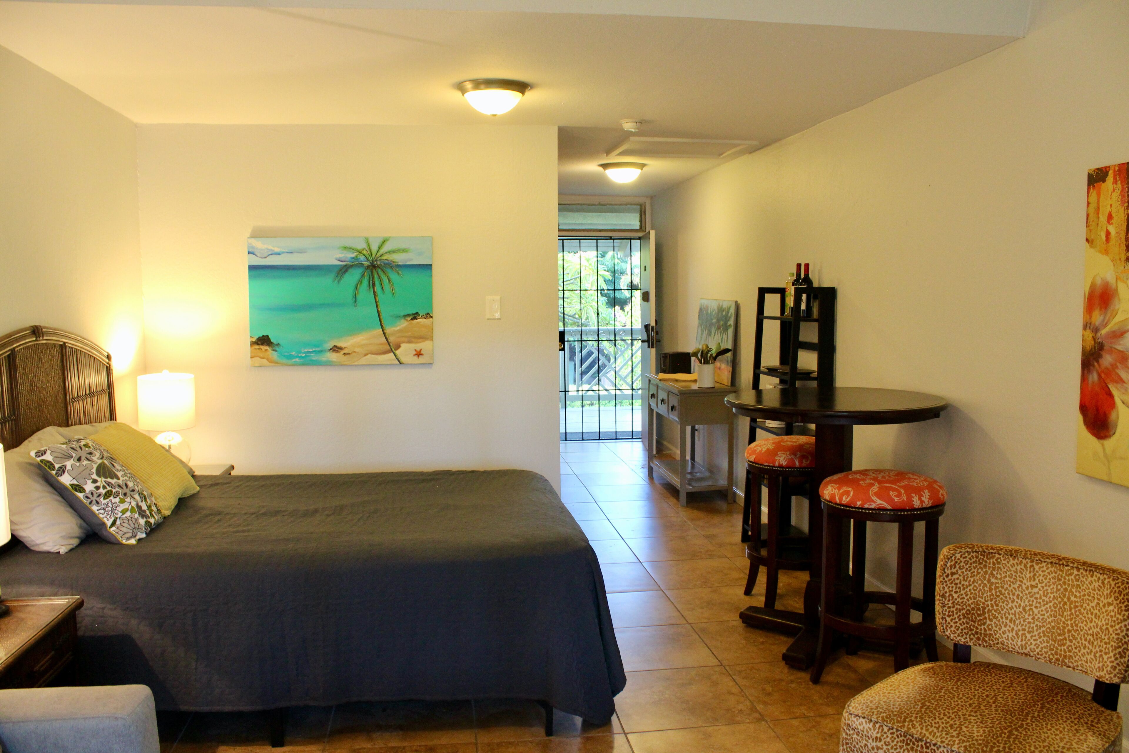 A bedroom with a bed, a desk, two chairs, and a painting of a beach scene.