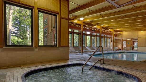 Una amplia piscina interior con ventanas transparentes, vigas de madera y suelo de baldosas.