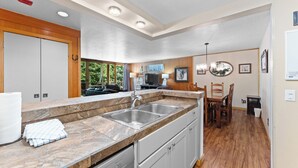 Una cocina moderna con encimera de mármol, pileta de acero inoxidable y muebles de cocina blancos. Hay una zona de comedor con sillas de madera y una mesa, y un televisor pequeño montado en la pared.