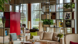 A modern living room with a sofa, chairs, a coffee table, and a bookshelf filled with books and decorative items. -  Intercityhotel Amsterdam Schiphol Airport (Hoofddorp)
