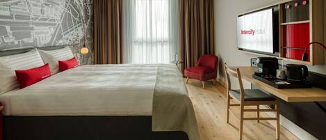 Una habitación de hotel moderna con una cama grande, un escritorio con cafetera, una silla roja y una pantalla de televisión grande que muestra el nombre del hotel.