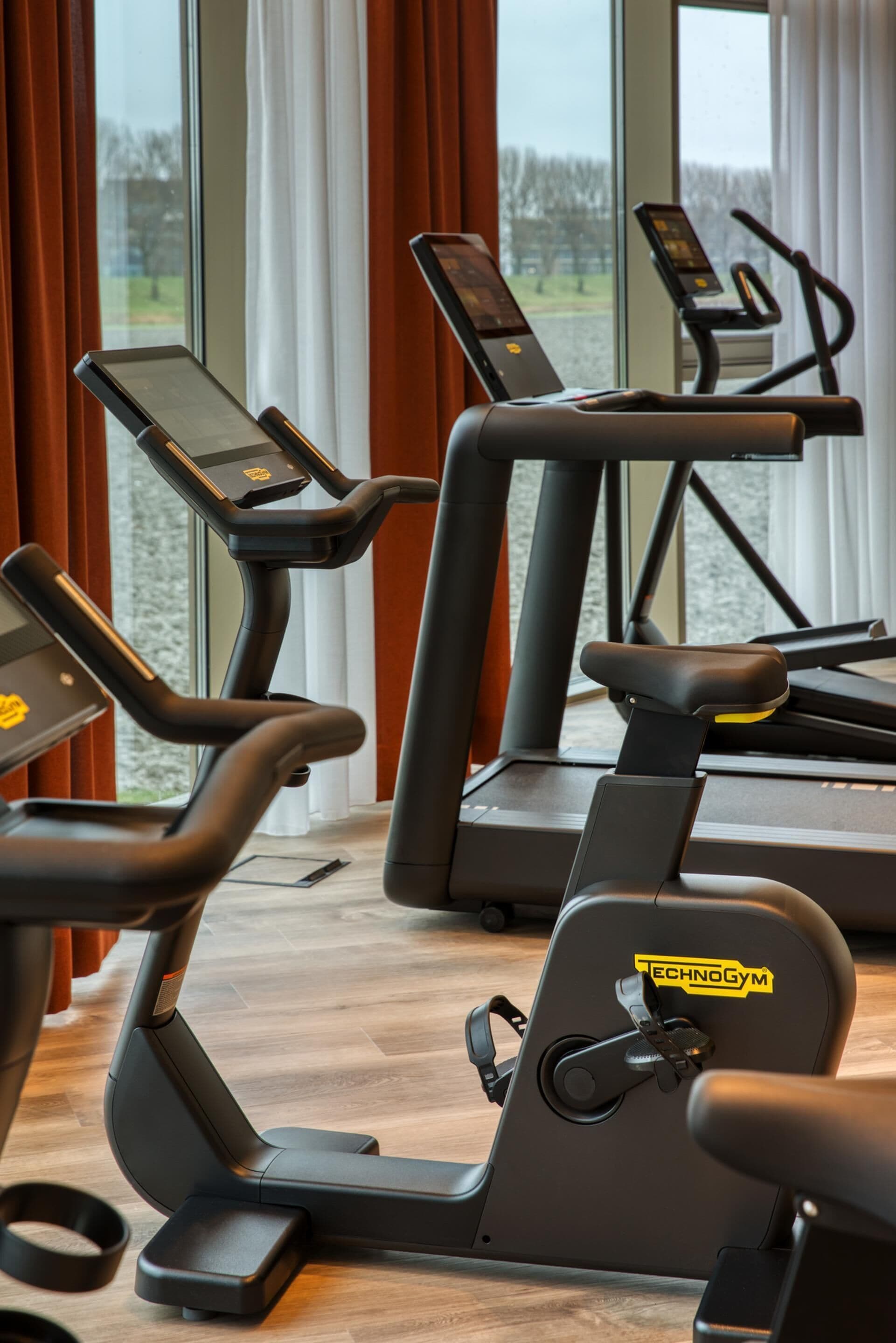 A gym with stationary bikes and a treadmill, large windows, and a view of trees.