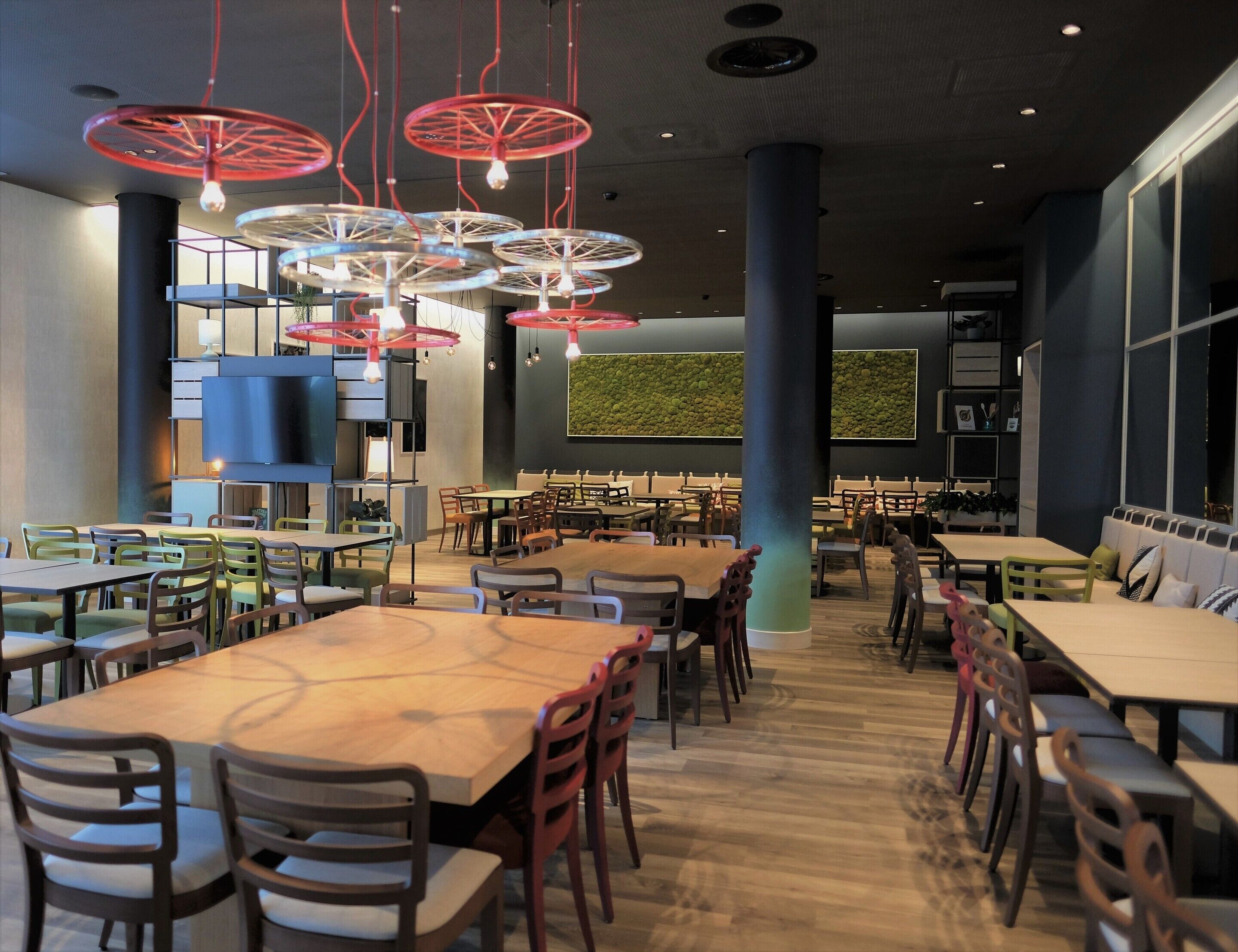 Un intérieur de restaurant moderne, avec de nombreuses tables et chaises, des suspensions rouges et blanches, et un grand mur vert orné d’un motif texturé.