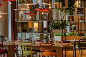 A restaurant with wooden tables and chairs, hanging pendant lights, and shelves with plants and books. -  Intercityhotel Amsterdam Schiphol Airport (Hoofddorp)