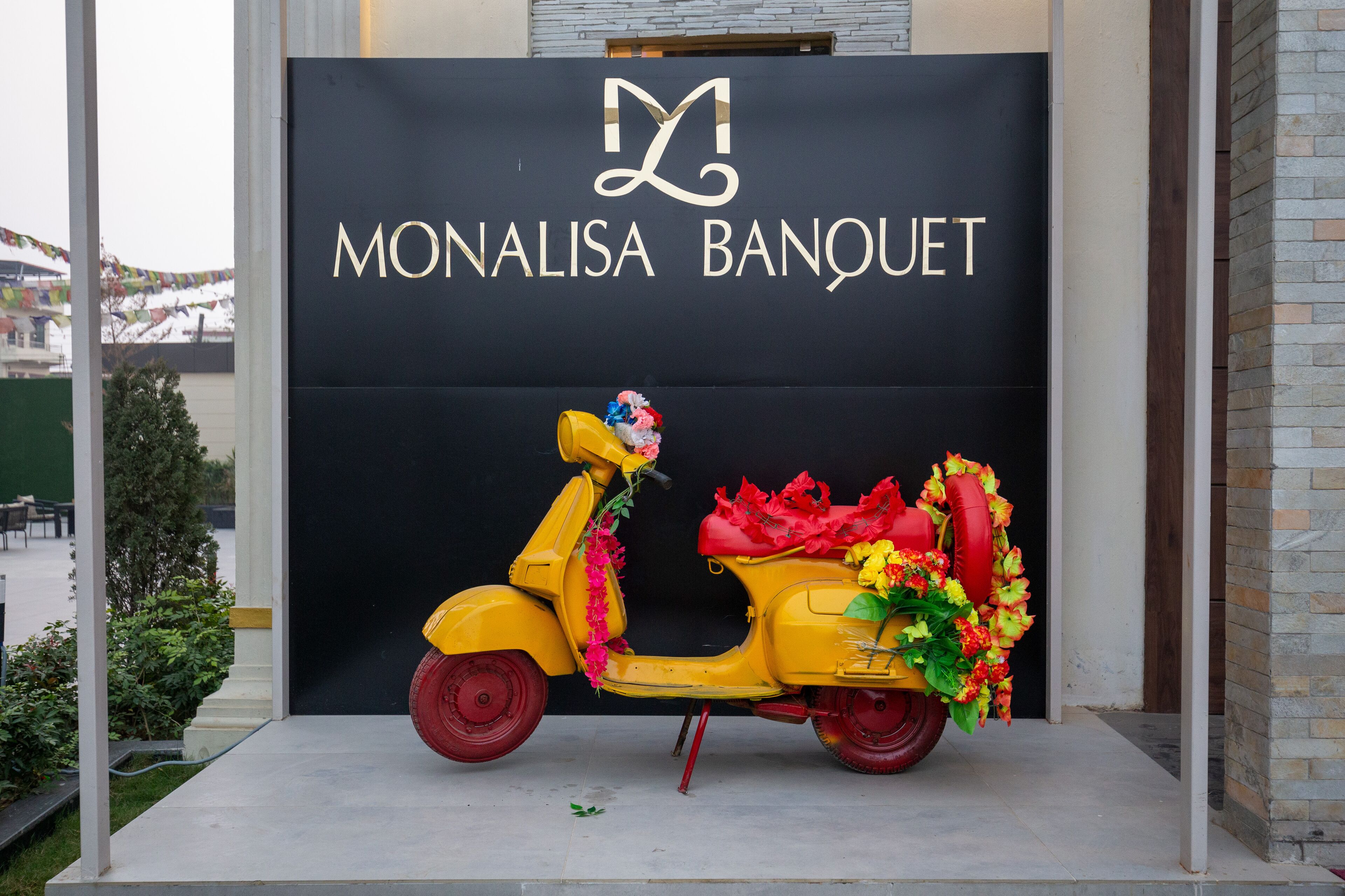 Un patinete amarillo adornado con flores y cintas frente a un letrero negro que dice 'MONALISA BANQUET'.