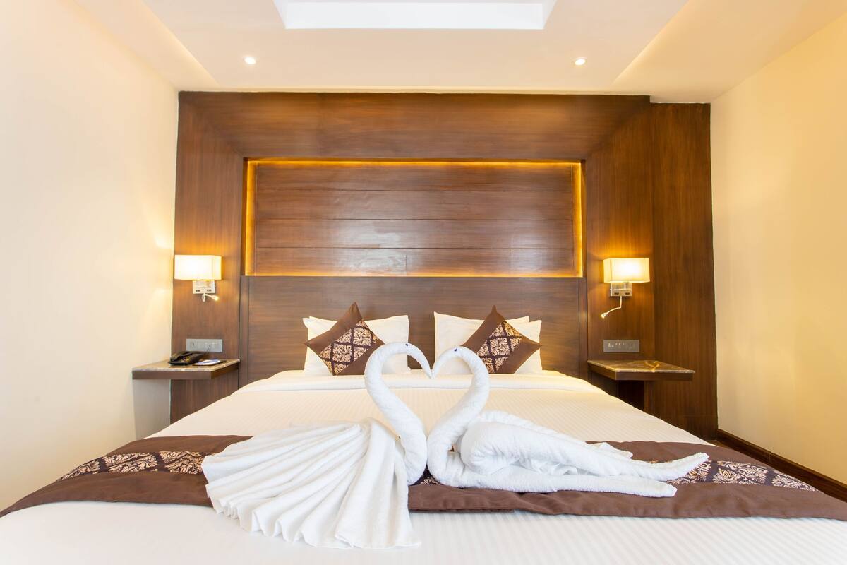 A hotel room with a large bed, two bedside lamps, a wooden headboard, and a heart-shaped towel arrangement.
