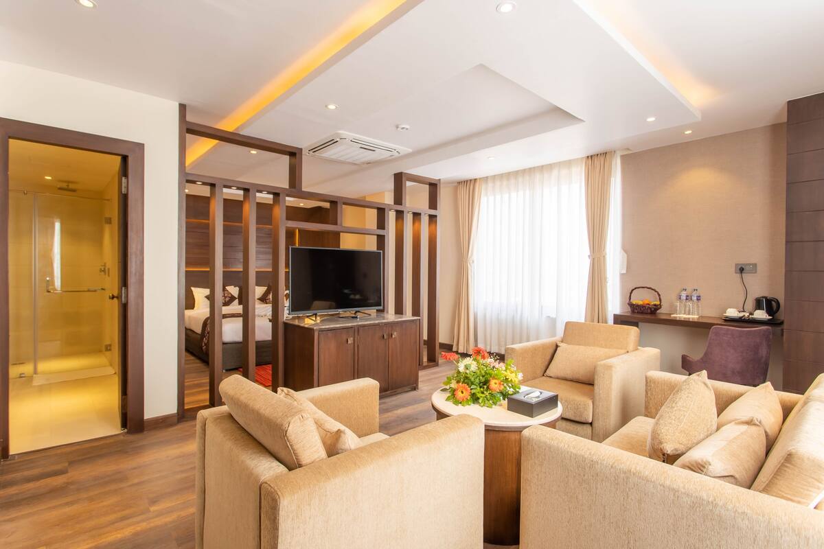 A modern hotel room with a living area featuring a sofa set, a coffee table with flowers, a flat-screen TV, and a wooden cabinet.