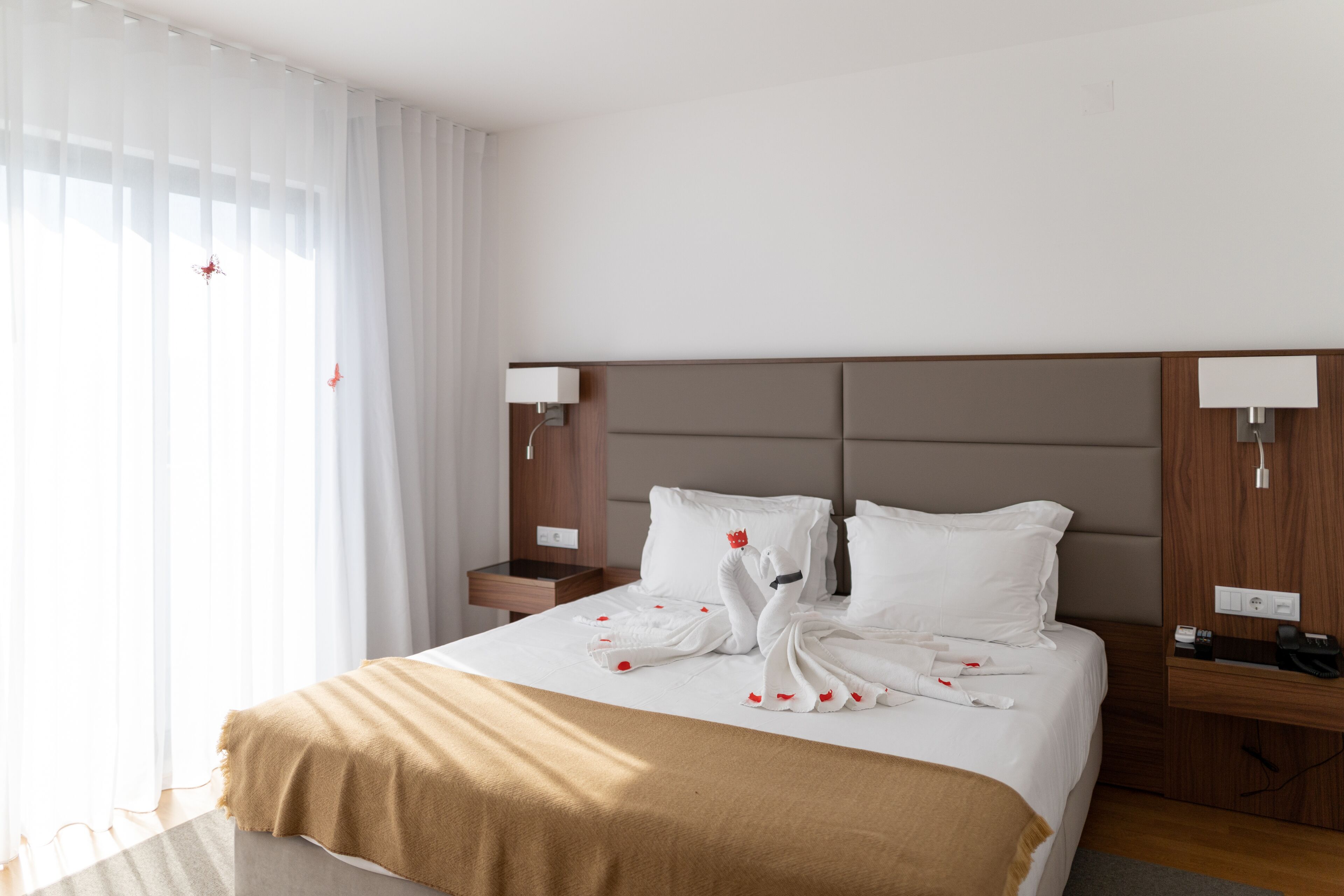 Habitación de hotel con cama, cortinas blancas, cabecera de madera, lámparas de noche y una manta marrón.