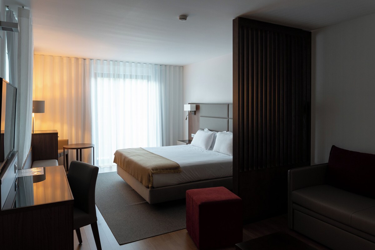 Quarto de hotel com cama, escrivaninha, cadeira, sofá e televisão.