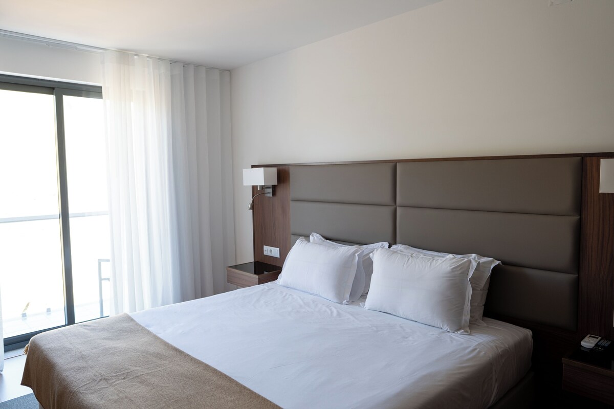 Quarto de hotel com uma cama grande, uma mesa de cabeceira, uma luminária e vista para o exterior.