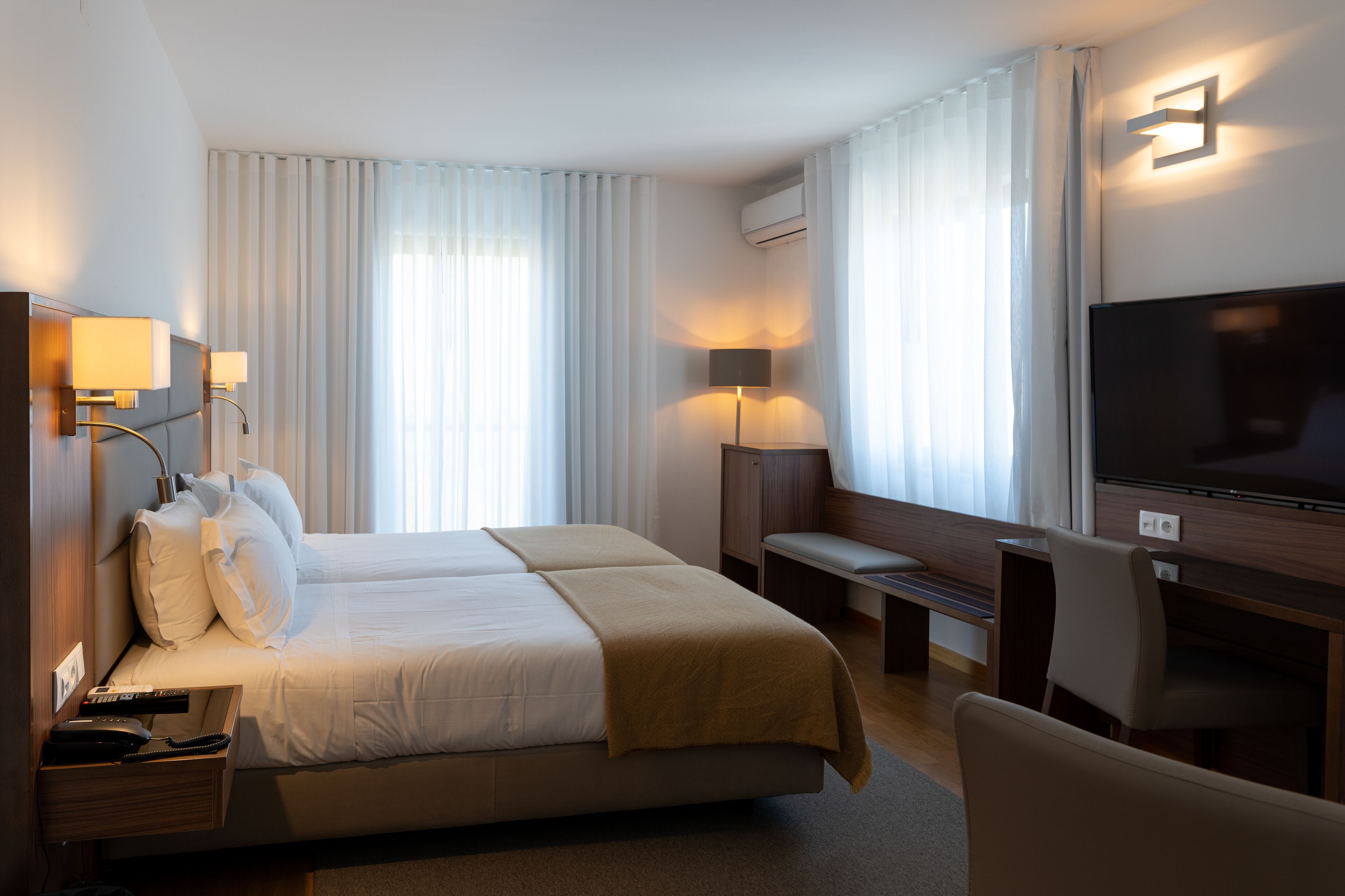 A hotel room with a bed, bedside tables, a desk, a chair, a TV, and curtains.