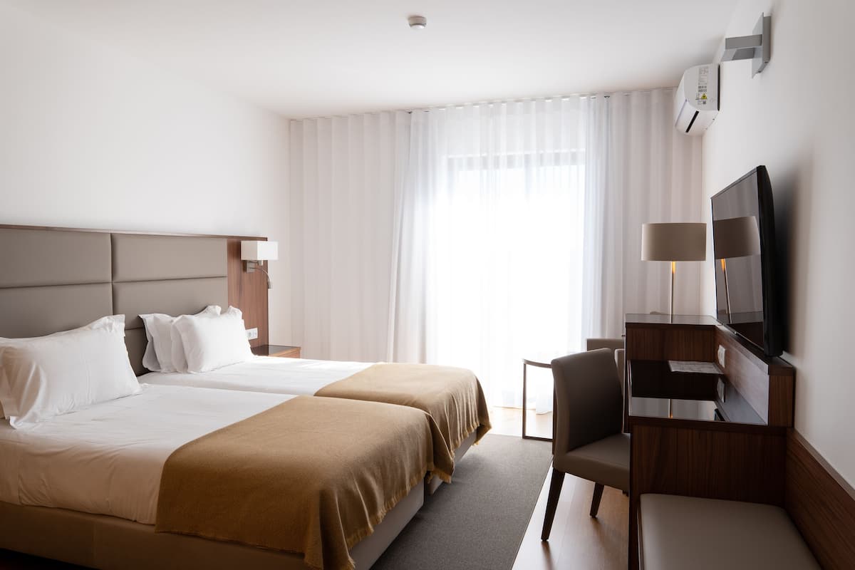 A hotel room with two beds, a desk with a chair, a television, and an air conditioning unit.
