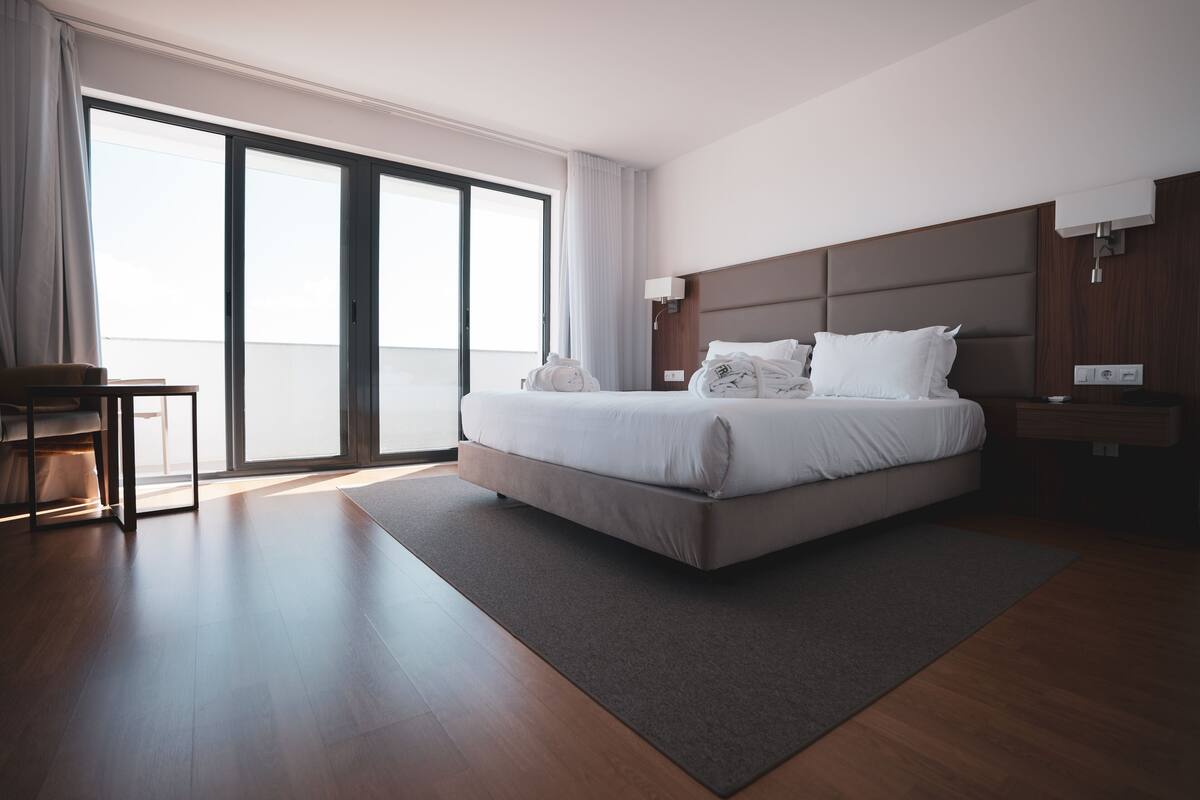 A modern hotel room with a large bed, a desk, and a view of the outdoors through a sliding door.