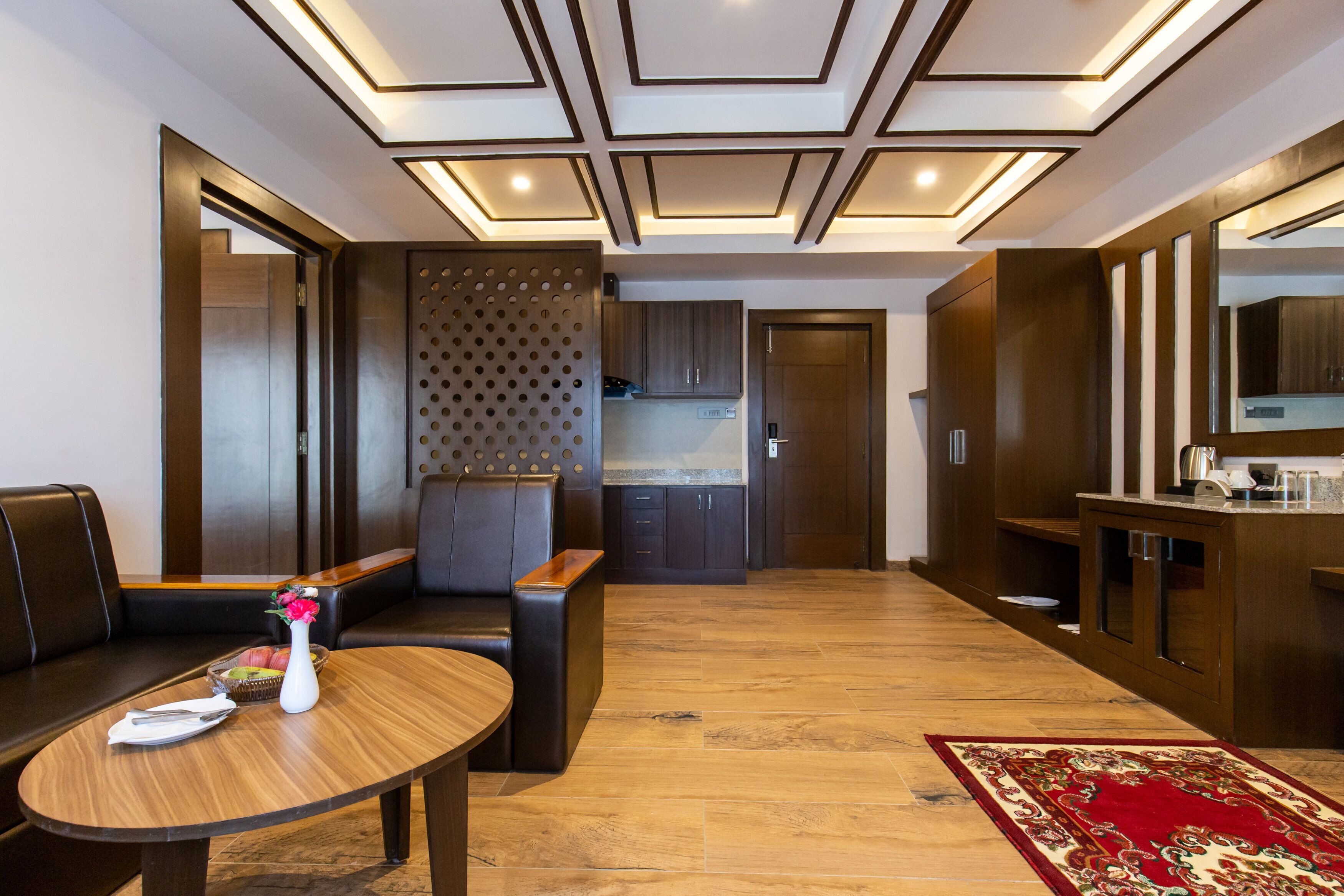 A modern hotel room with a dining area, a sofa, a table with a vase, and a kitchen area.