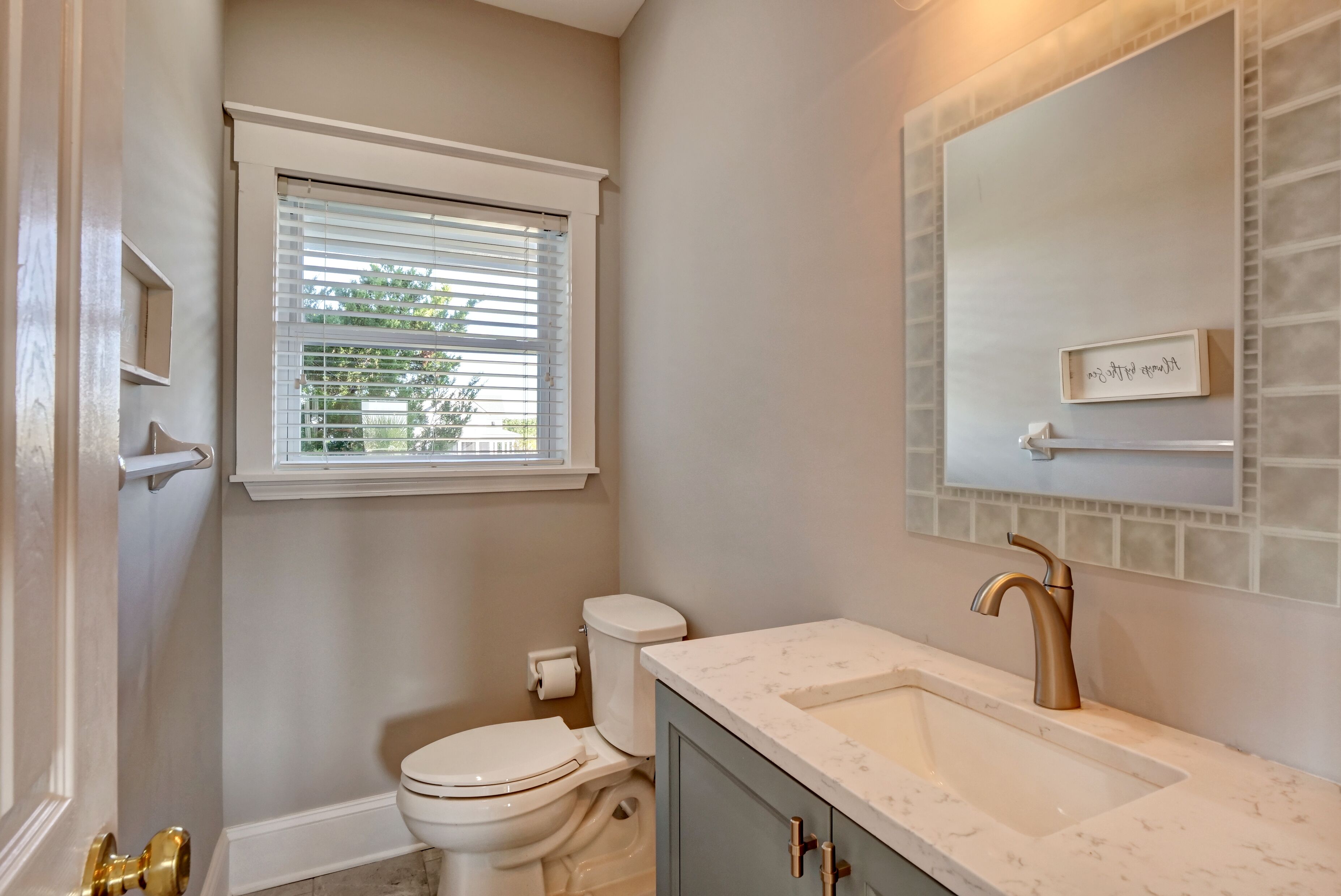 A bathroom with a toilet, sink, vanity, window, and a mirror with a nameplate.