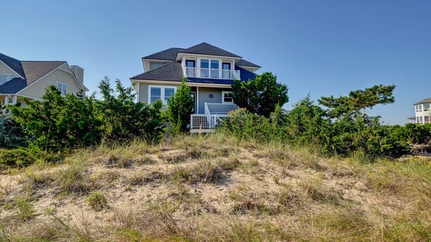 A two-story house with a balcony, surrounded by dense shrubbery and sandy terrain. - Bald Head Island. Oceanfront 2nd Row. 4 bedrooms. 3.5 baths. BHI Club access. (Bald Head Island)