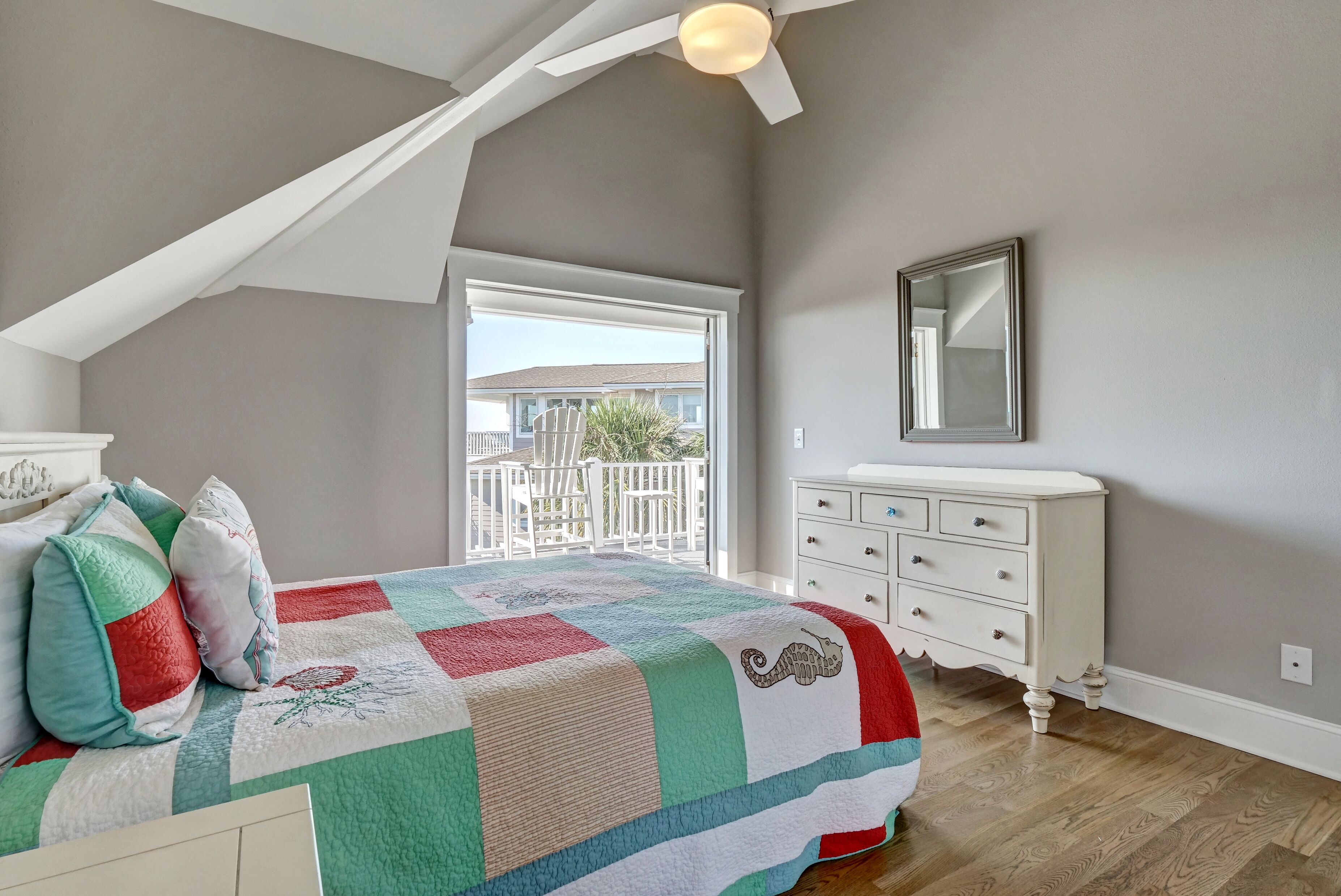 A bedroom with a sloped ceiling, a large window, a bed with a colorful quilt, a dresser, and a mirror on the wall.