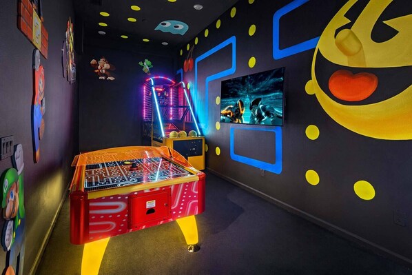 Una habitación con mesa de fútbol de mesa, una máquina recreativa iluminada con neón y una pantalla de televisión mostrando un juego de carreras.