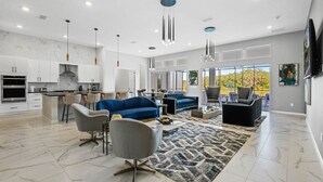 Una cocina moderna con armarios blancos y una isla central, una sala de estar con un sofá azul y sillones grises, y un comedor con una mesa y sillas.