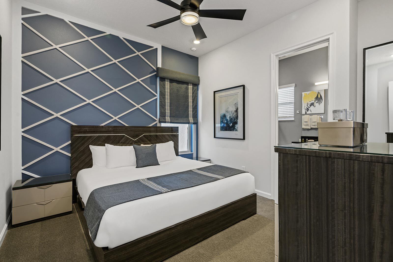 Un dormitorio moderno con cama, mesita de noche, cómoda, ventilador de techo y baño con espejo y lavabo.