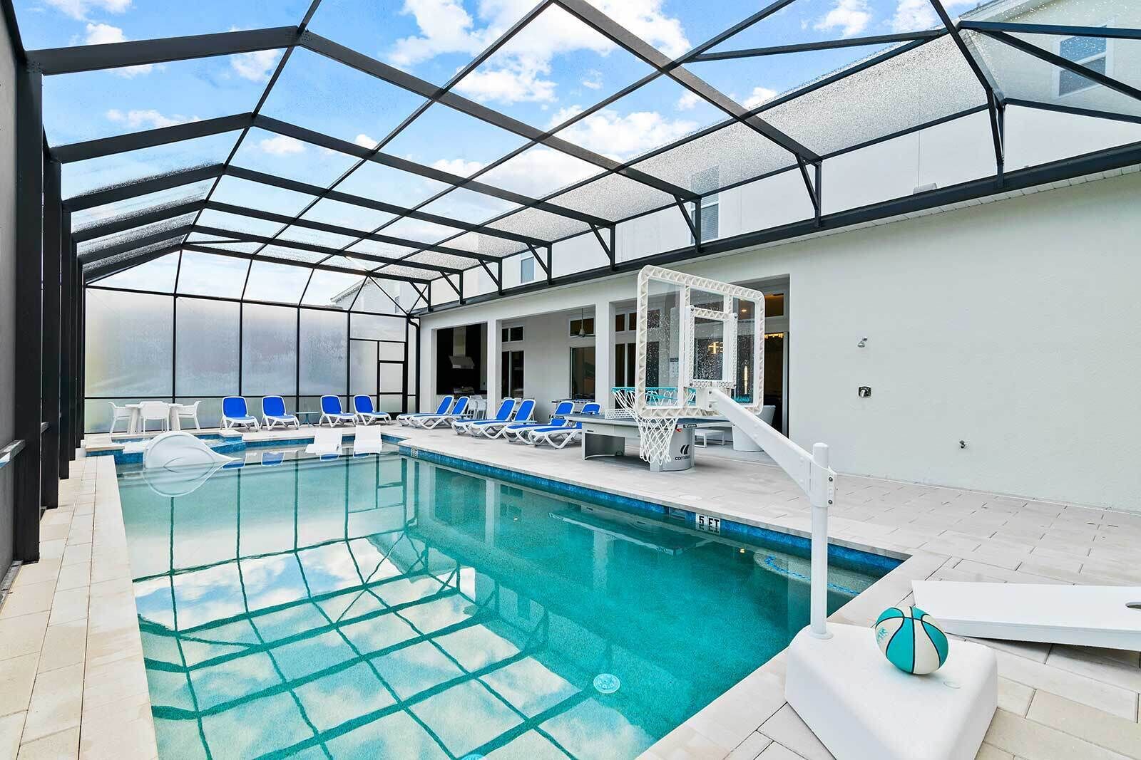 An indoor swimming pool with clear glass walls, blue lounge chairs, and a basketball hoop.