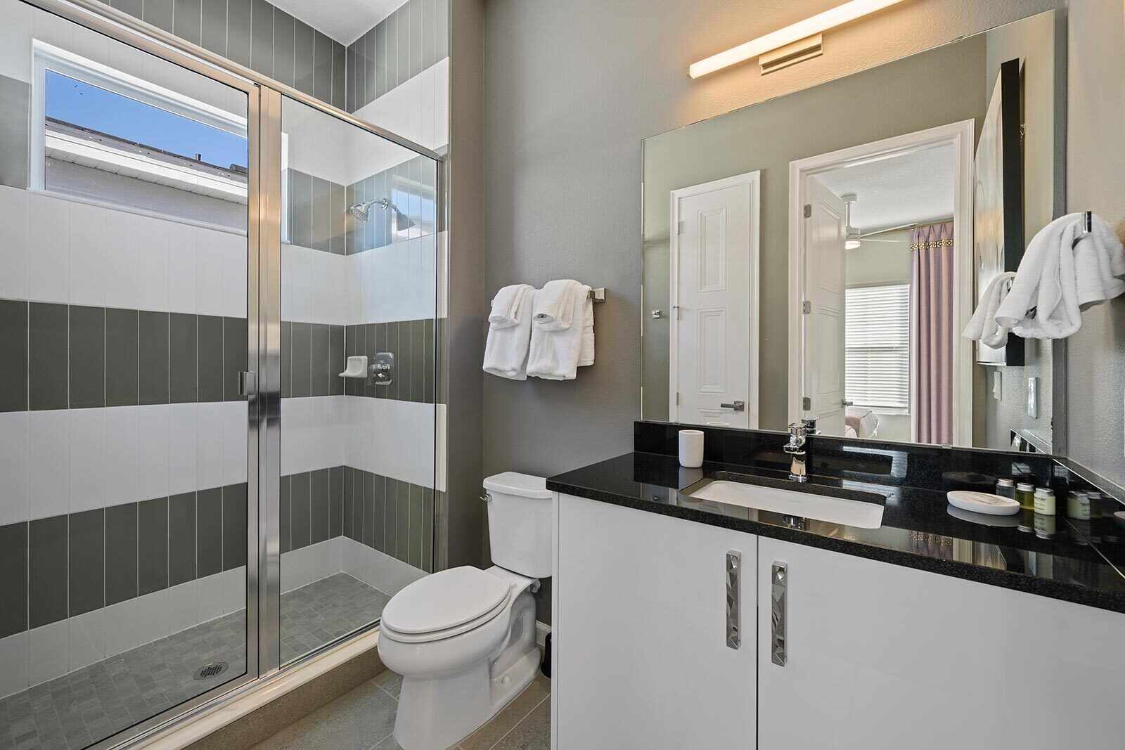 Une salle de bain moderne avec une douche à l’italienne en verre, des toilettes blanches, un lavabo noir et un grand miroir.