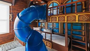 A room with a large blue spiral slide, wooden walls, and a bed with a view of the sea through arched windows.
