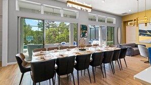 A modern dining area with a long table set for a meal, large windows offering a view of the outdoors, and contemporary furniture.