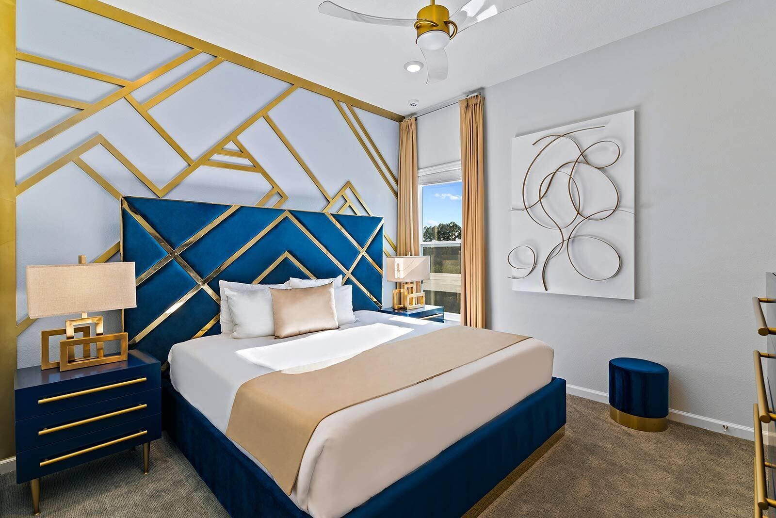 Une chambre moderne avec une tête de lit à motifs géométriques, un grand lit recouvert d’une couverture beige et bleue, une table de chevet avec une lampe et une œuvre d’art abstraite accrochée au mur.