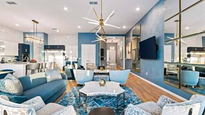 A modern living room with a blue sofa set, a coffee table, and patterned chairs. A large mirror and a chandelier are visible. - Super Villa Near Disney w Sci-Fi Arcade, Space Theme BR, Theater, Gym, Spa Pool (Davenport)