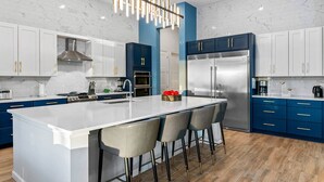 A modern kitchen with a large island, white cabinets, blue accents, stainless steel appliances, and a unique pendant light. - Super Villa Near Disney w Sci-Fi Arcade, Space Theme BR, Theater, Gym, Spa Pool (Davenport)