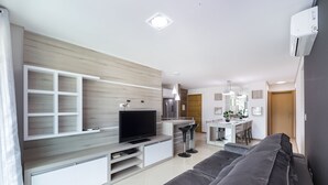 A modern living room with a TV, built-in shelves, a grey sofa, and a kitchen area in the background.