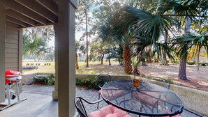 A covered patio with a round table and chairs, a grill, and a view of a landscaped garden with trees and a lawn. - Beautifully Updated One Bedroom Villa Great for a Couples Getaway! (Hilton Head)