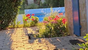 Un camino pavimentado que lleva a una casa con puerta azul, rodeada de plantas en flor y una zona de piscina cubierta.
