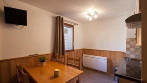 Et lille køkken med et træbord, stole, en potteplante, et tændt stearinlys og et fjernsyn ophængt på væggen.