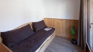 Et lille, hyggeligt rum med en sengebænk i træ, en enkelt stueplante og et vindue med gardiner.