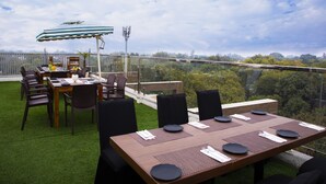 Un área de comedor al aire libre con vista a un paisaje urbano, una mesa preparada con platos y cubiertos, y un paraguas a rayas.