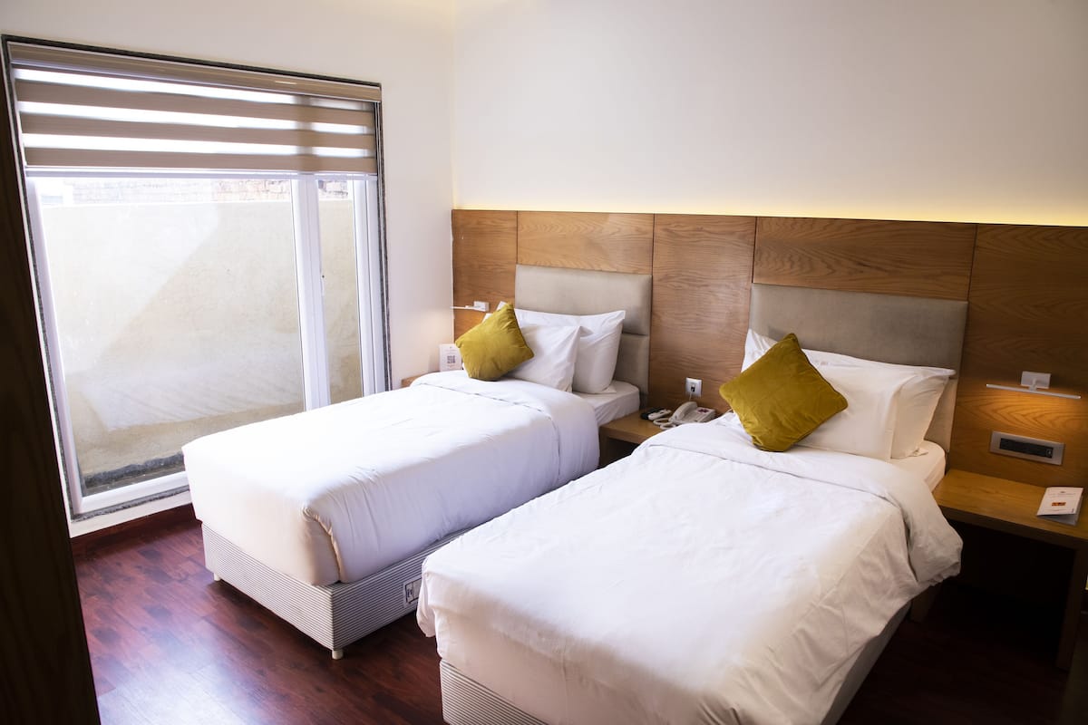 Une chambre d’hôtel avec deux lits, une tête de lit en bois, une table de chevet avec un téléphone et une fenêtre avec des stores.