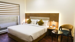 Habitación de hotel con una cama grande, un cabecero de madera, una mesita de noche con lámpara, una silla y una ventana con persianas.