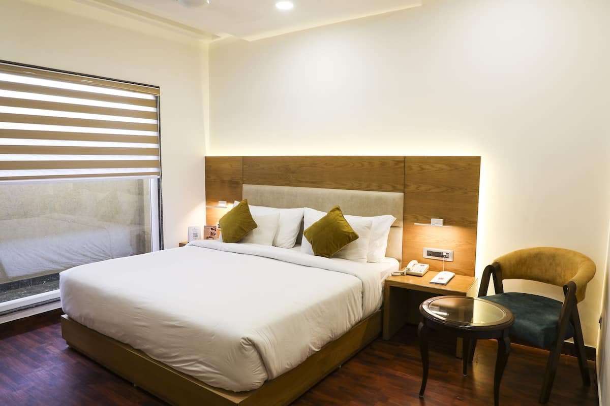 Une chambre d’hôtel avec un grand lit, une tête de lit en bois, une table de chevet avec une lampe, une chaise et une fenêtre avec des stores.