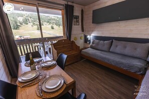A cozy room with a wooden dining table set for two, a sofa, and a large window offering a view of a forested area.