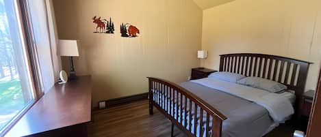 A bedroom with a wooden bed, a nightstand, a desk with a lamp, and a window.